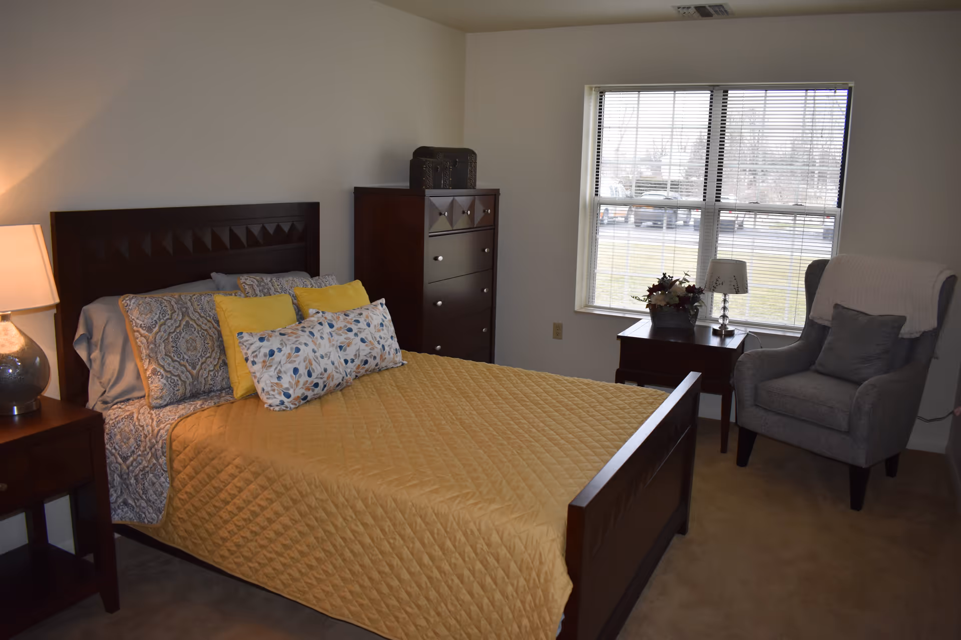 A neatly made bed with a yellow quilt and multiple pillows in a bedroom. The room features a wooden headboard, a nightstand with a lamp, a tall dresser, a window with blinds, a small table with a lamp and flower arrangement, and a gray armchair with a pillow and a throw blanket.