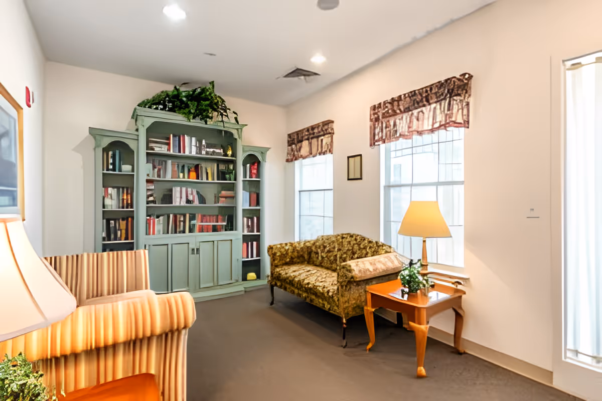 A cozy sitting room with upholstered chairs, a green bookcase filled with books, a floral settee, and a side table with a lamp under two windows.