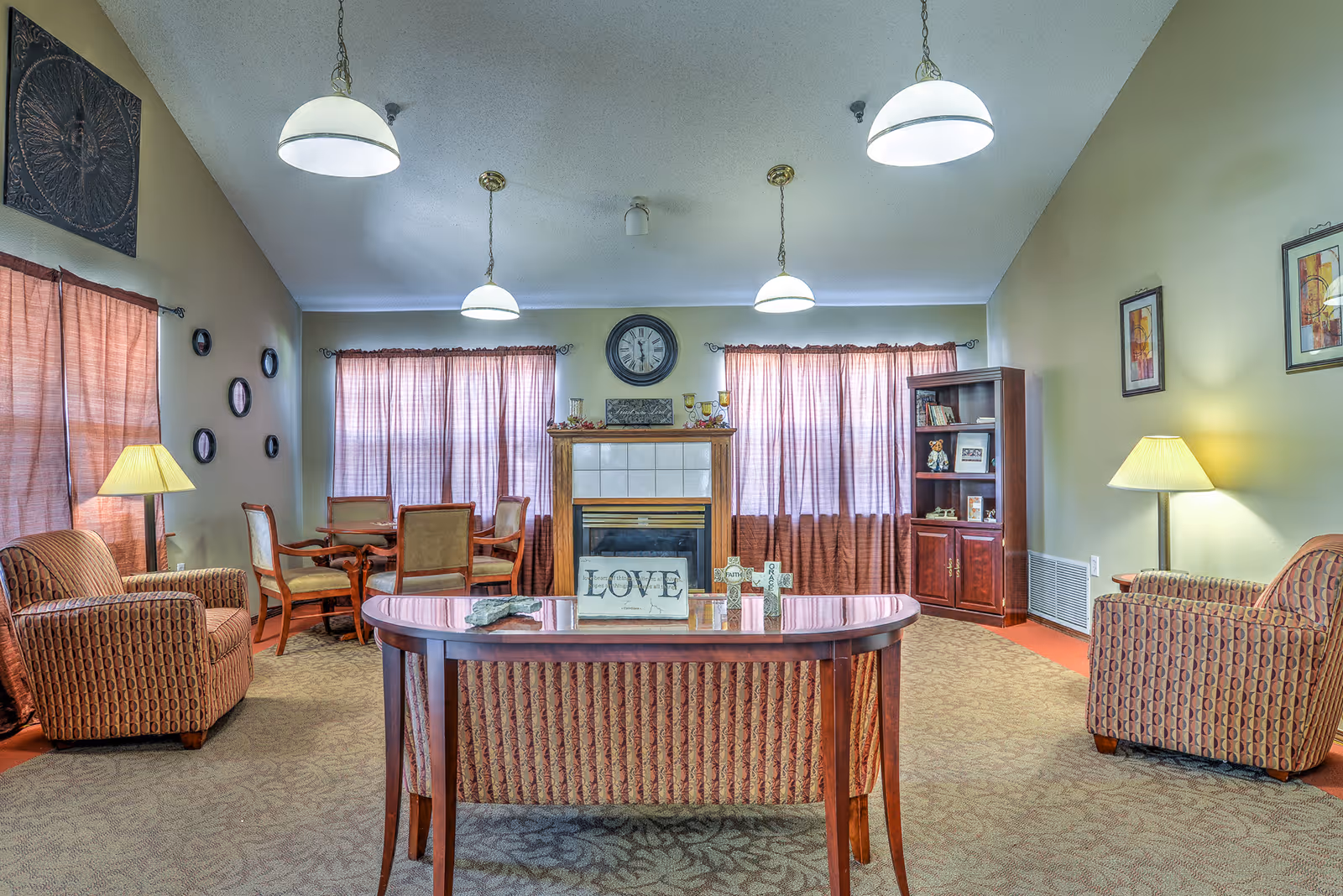 A cozy common room with upholstered armchairs, a central table, a fireplace and bookshelf, lit by pendant lights and lamps.