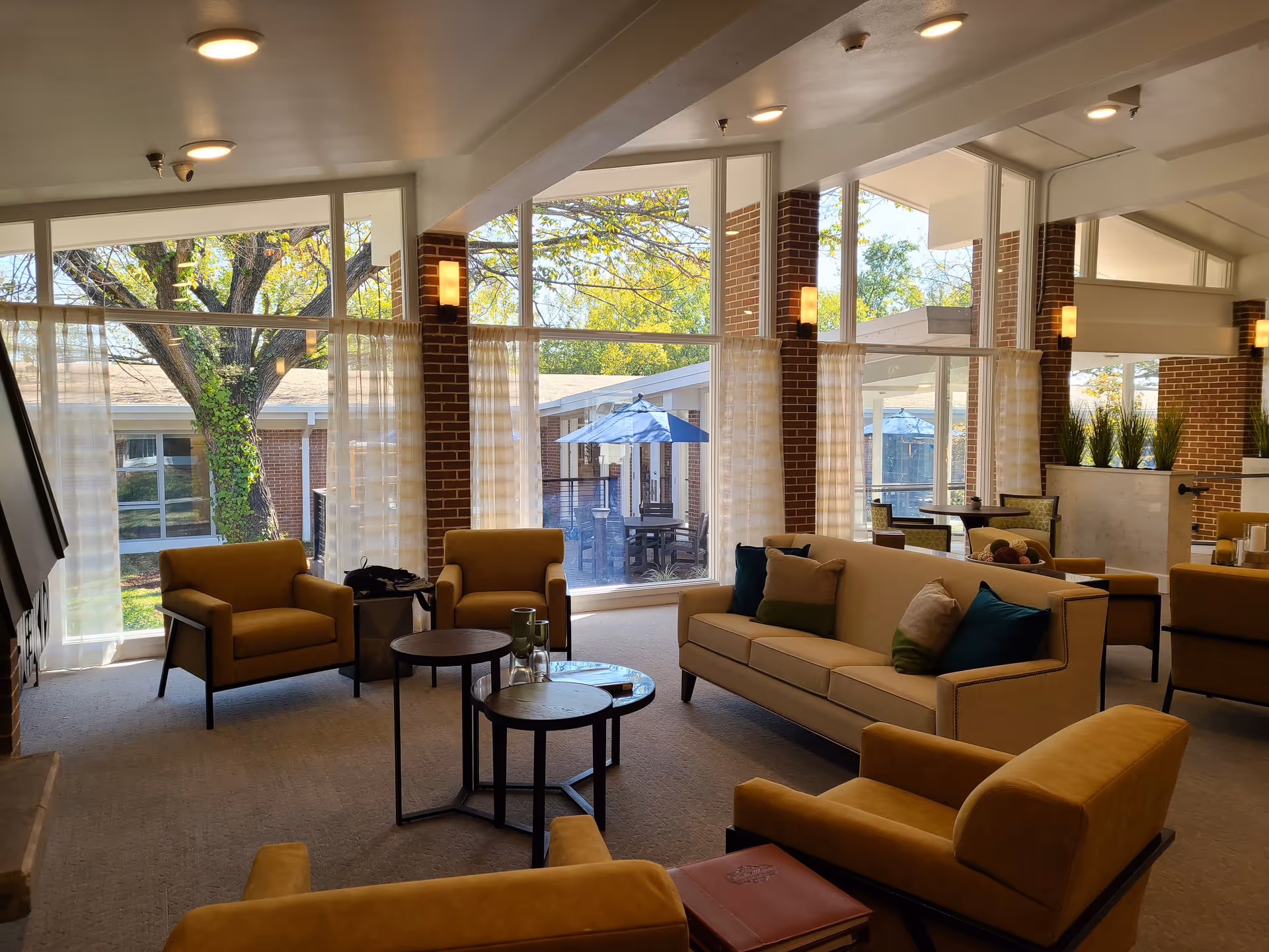 Bright common lounge with sofas and armchairs around coffee tables and large floor-to-ceiling windows overlooking a courtyard with an umbrella.