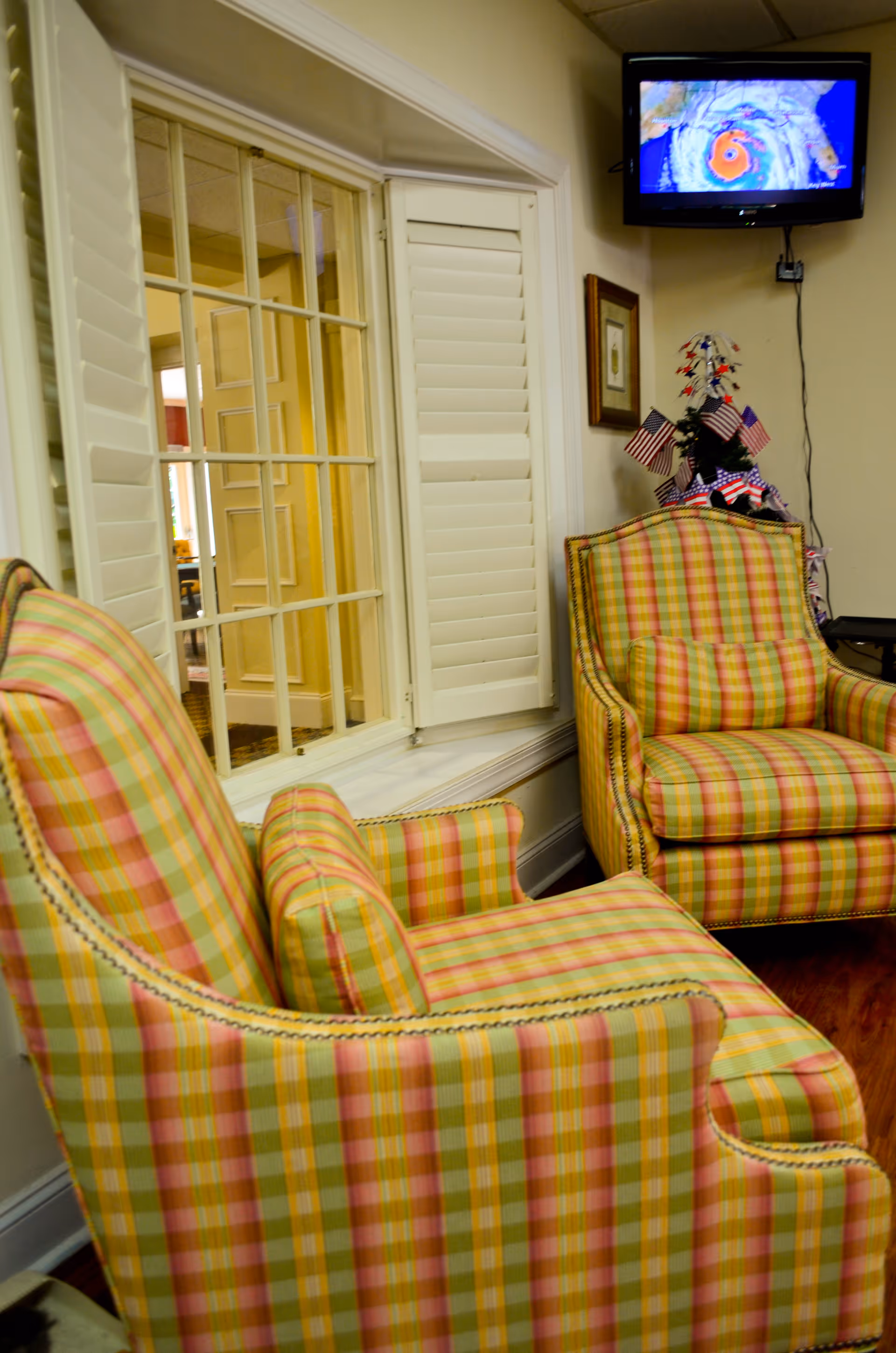 Two plaid upholstered armchairs in shades of yellow, green, and pink are placed in a cozy interior room with a window featuring white shutters. A small decorated tree with American flags is visible in the corner, and a wall-mounted TV displays a weather map with a hurricane image.