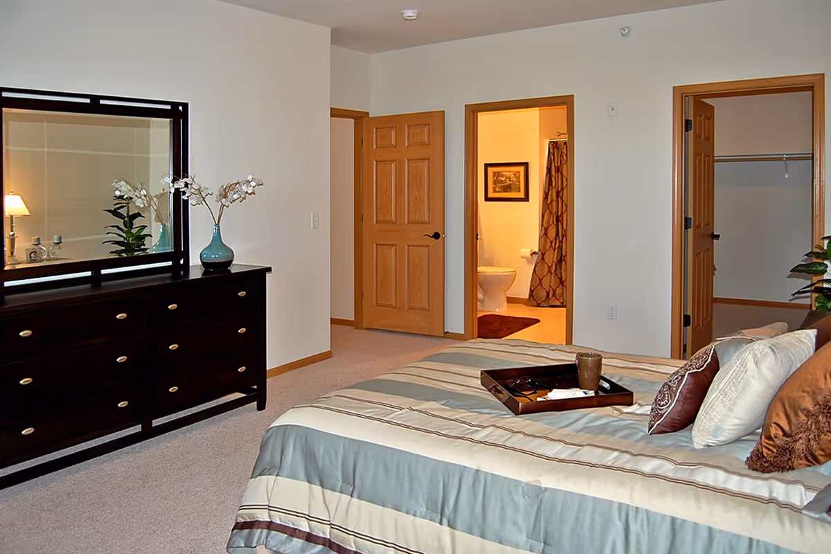 Well-lit bedroom with a bed and dresser, and open doors leading to a bathroom and a closet.