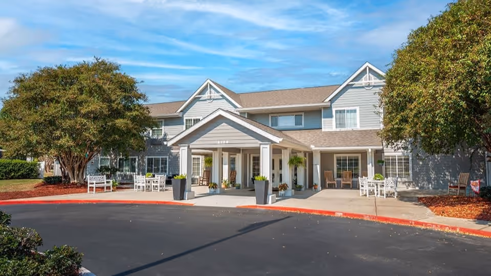 Two-story light-gray senior living facility with a covered porte-cochère entrance, outdoor seating, and landscaped trees.