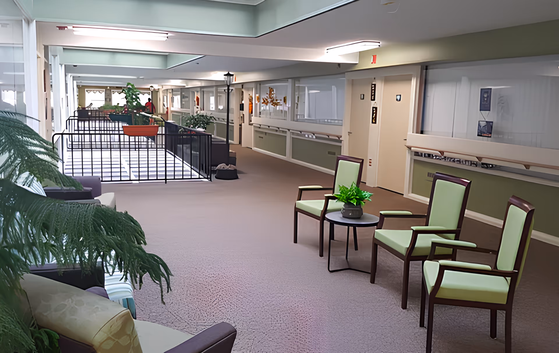 A spacious indoor hallway area in a senior living facility with several green cushioned chairs arranged around a small round table with a potted plant. The hallway has beige carpeting, green and white walls, large windows, and various plants placed along the corridor. There are doors and handrails along the walls, and the ceiling has fluorescent lighting.