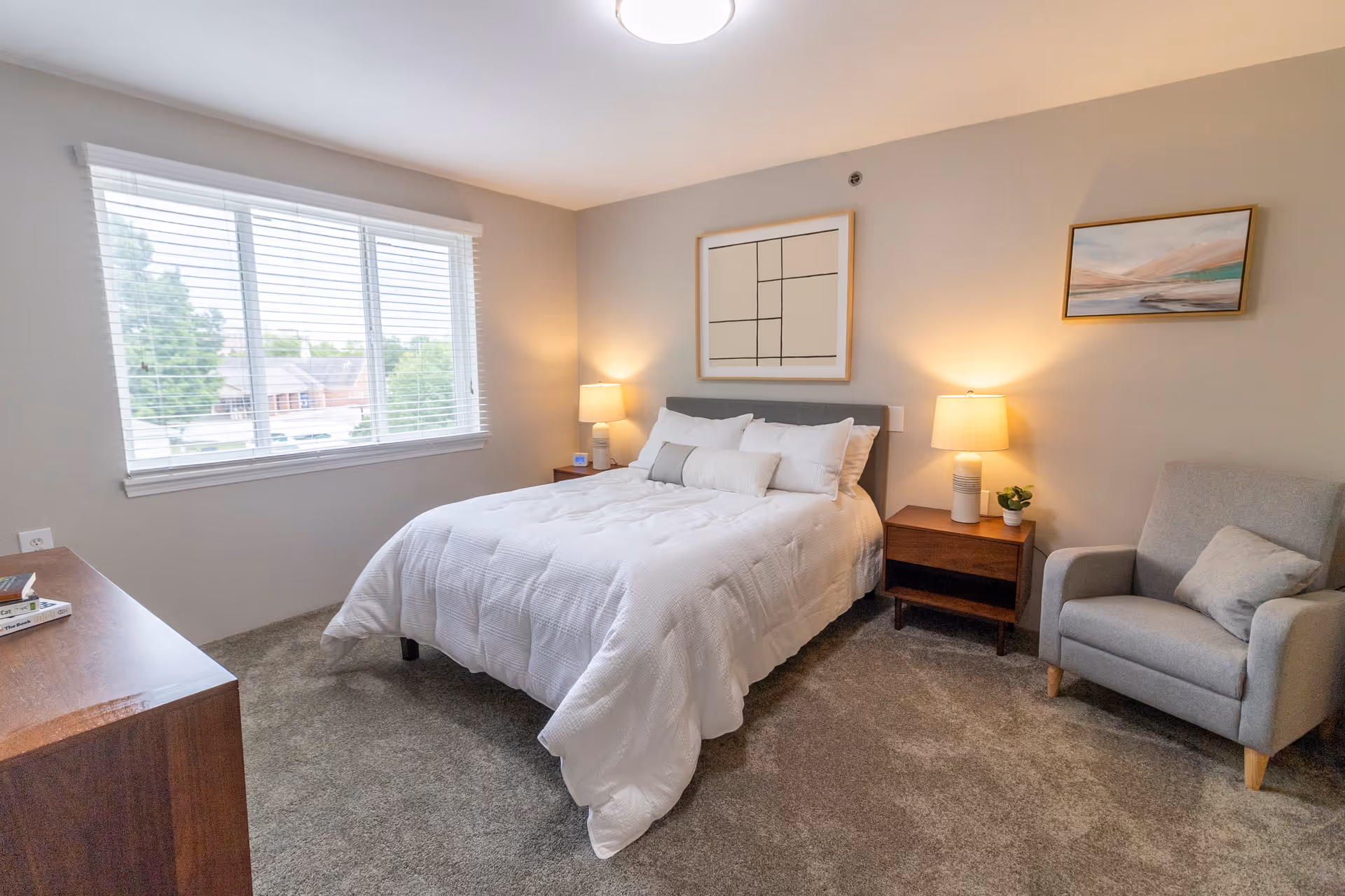 A bright and cozy bedroom with a large window letting in natural light. The room features a neatly made bed with white bedding and multiple pillows, two wooden nightstands each with a lamp, a gray armchair with a cushion, a wooden dresser, and two framed artworks on the walls. The floor is carpeted in a neutral tone.