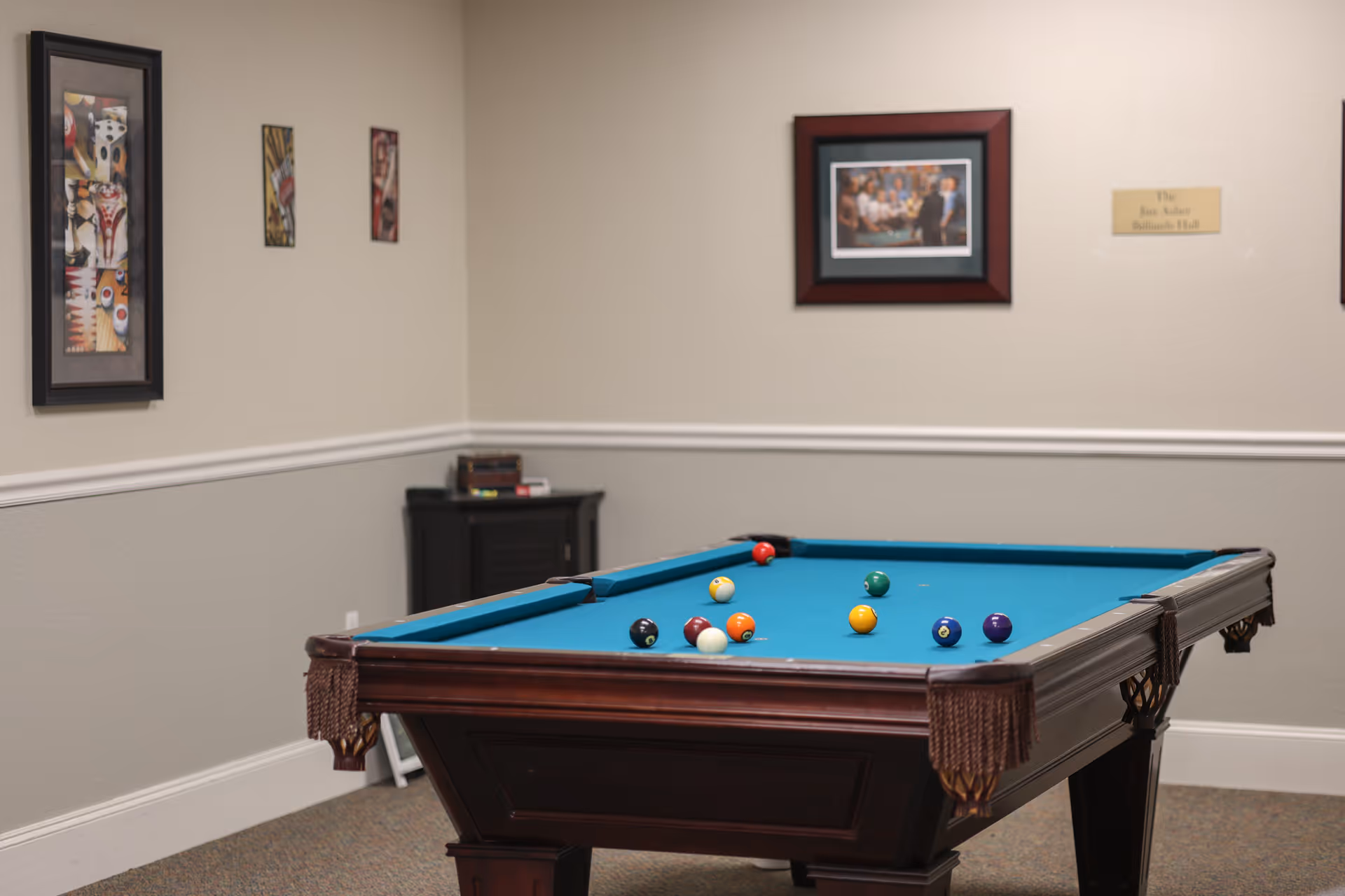A billiards hall with a pool table featuring a blue felt surface and several billiard balls scattered on it. The room has beige walls with white trim, framed artwork on the walls, and a small cabinet in the corner with books on top.