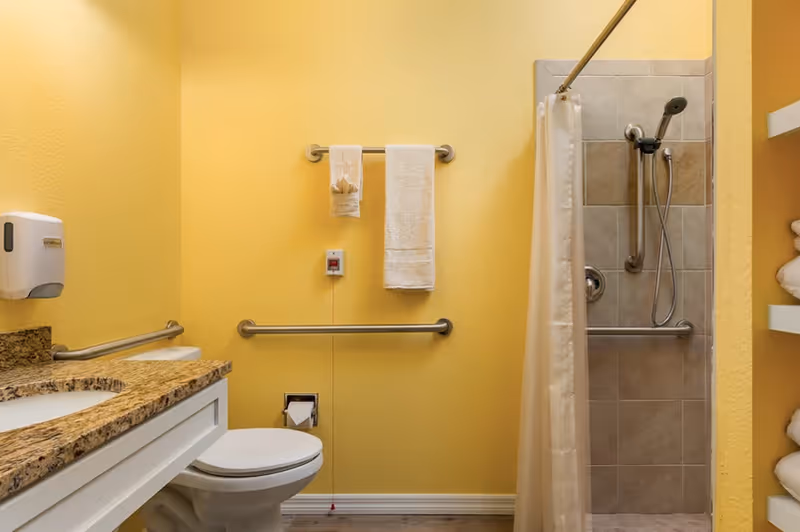 Accessible bathroom with yellow walls, a toilet, granite vanity, grab bars, and a tiled walk-in shower with a curtain.