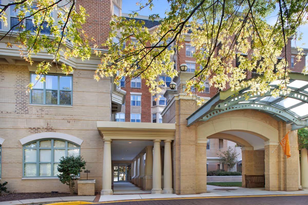 Exterior view of Brighton Gardens of Friendship Heights showing a covered entrance with columns, a brick and beige facade, windows, and a tree with green leaves in the foreground.