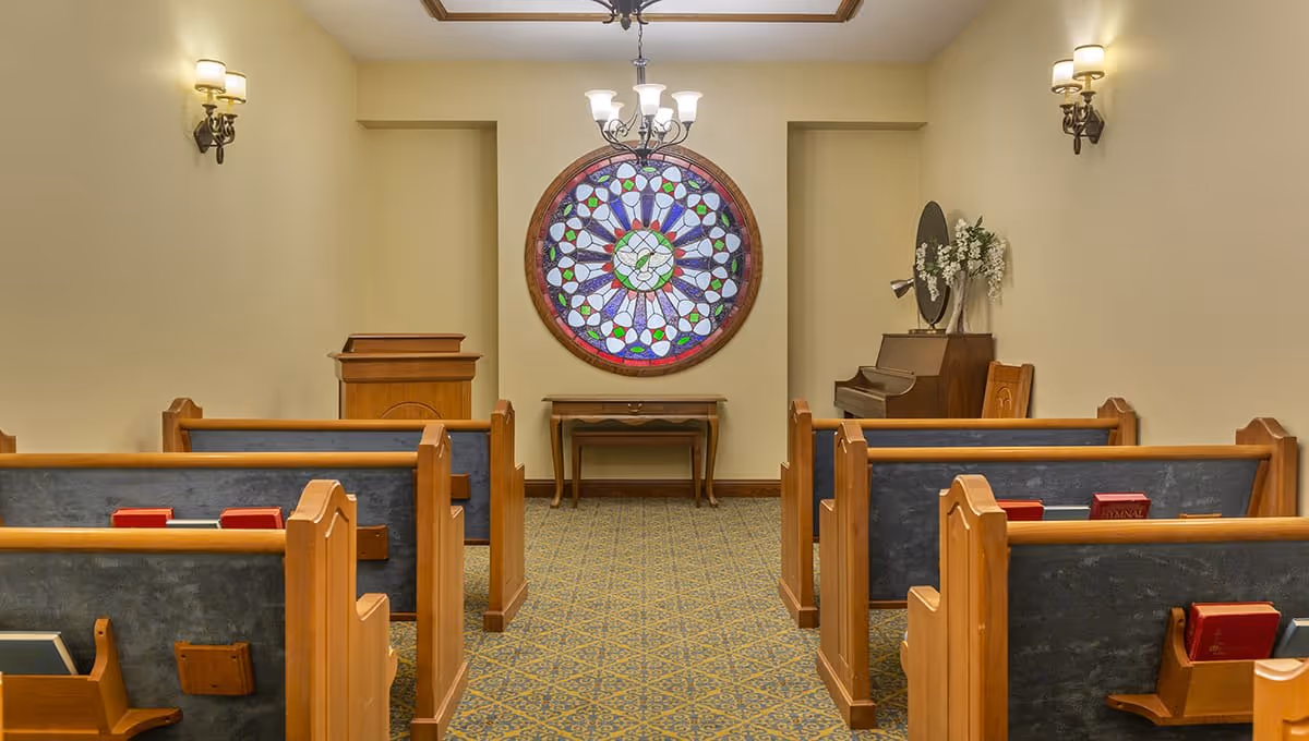 Interior view of a small chapel or worship room with wooden pews, a stained glass circular window on the far wall, a wooden podium, a piano with a flower vase on top, and wall-mounted light fixtures.