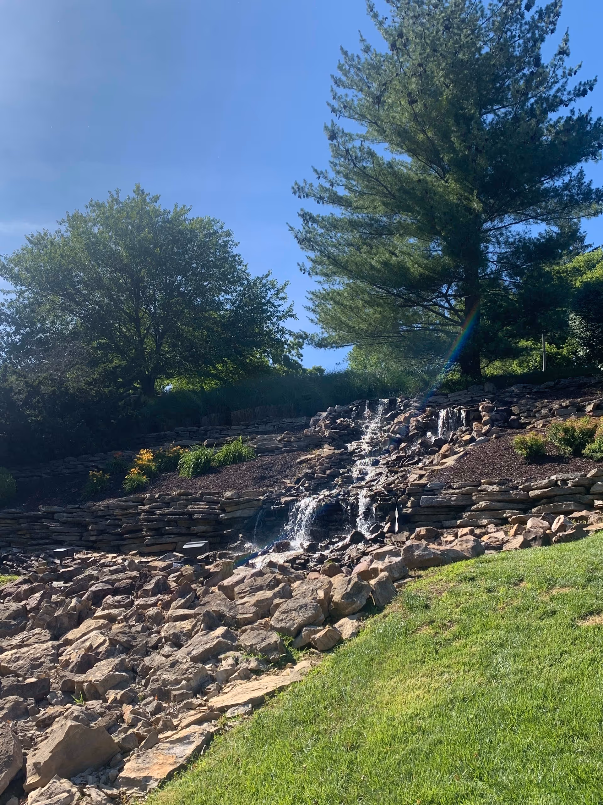 Outdoor scene featuring a landscaped rock waterfall surrounded by green grass, bushes, and tall trees under a clear blue sky.
