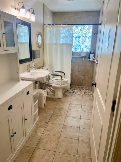 A clean, accessible bathroom with beige tiled floor and walls. The room features a white sink with a mirror and light fixture above it on the left side, a white toilet with arm supports, and a walk-in shower with a white curtain and a window with floral patterned privacy film. There is a white cabinet for storage on the left side and a white door on the right side of the image.