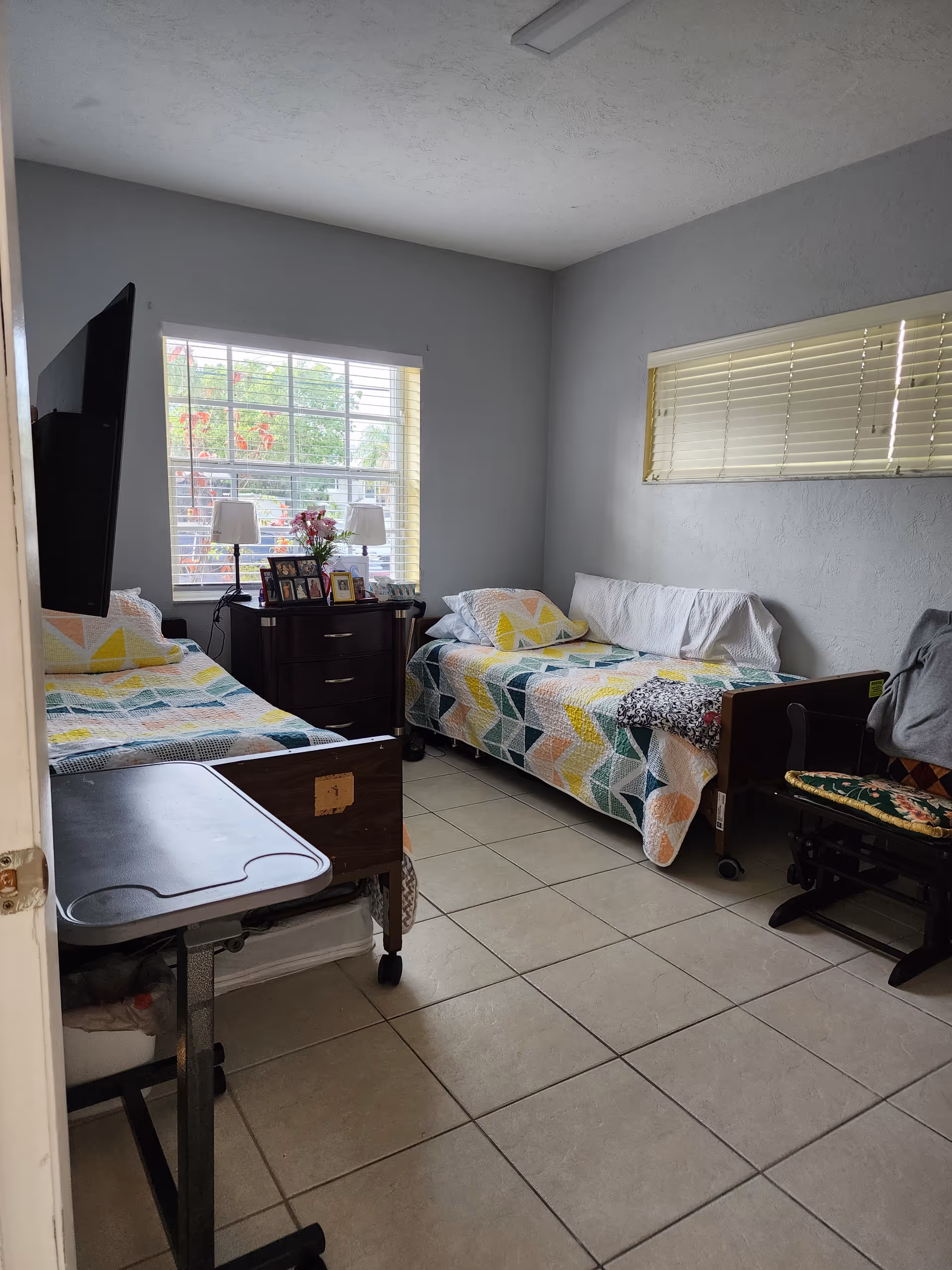 A small bedroom with two single beds on opposite walls, each covered with colorful geometric patterned quilts. Between the beds is a dark wooden nightstand with two lamps, framed photos, and a vase with flowers. A flat-screen TV is mounted on the wall near one bed, and a chair with a cushion is placed near the other bed. The room has tiled flooring and two windows with blinds, allowing natural light to enter.
