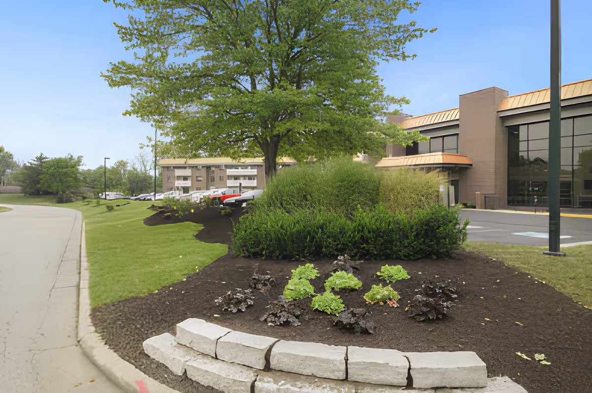 Landscaped driveway and entrance with planted beds, a large tree, and the front facade of a senior living building.