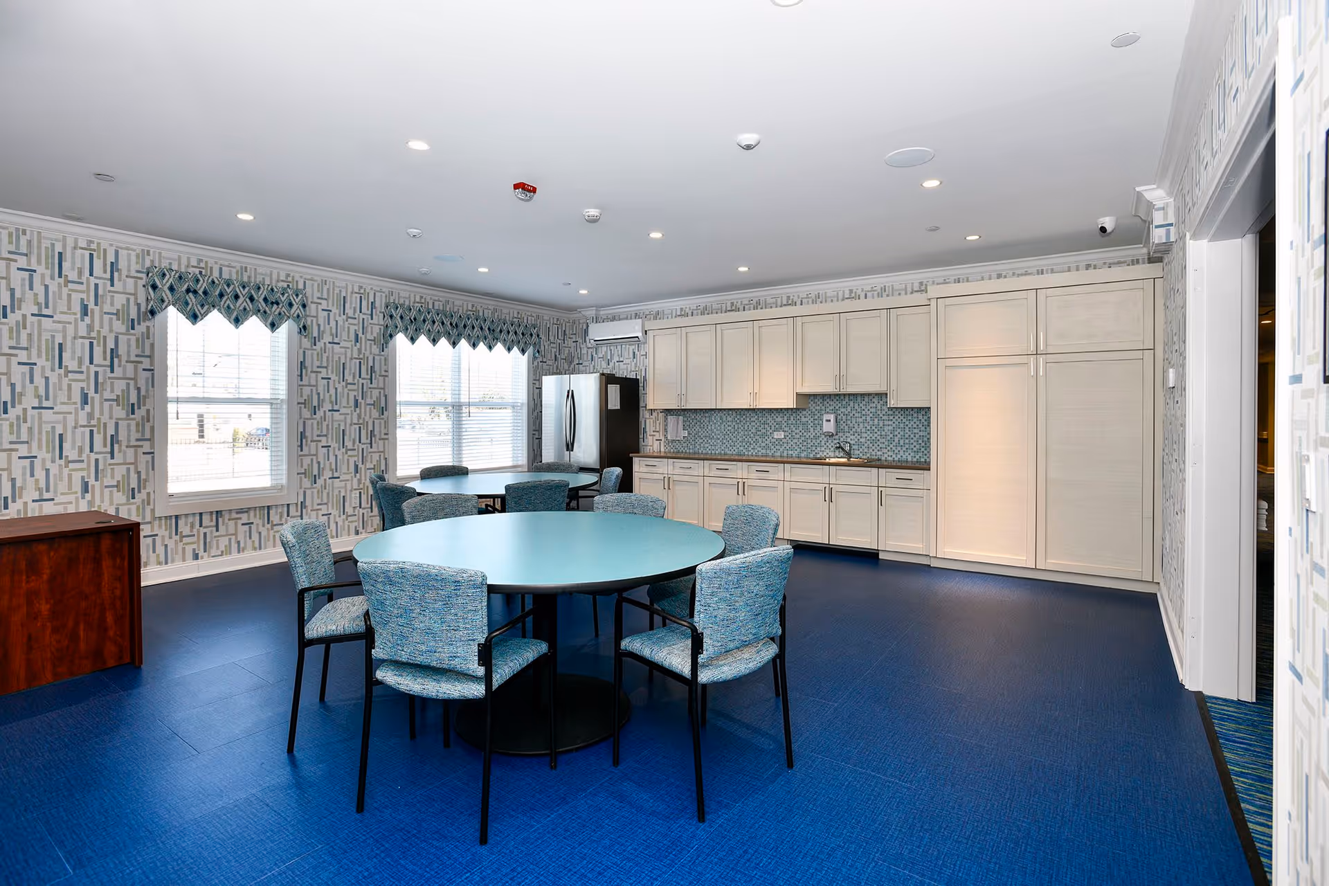 Bright communal dining area with round tables and upholstered chairs in front of a kitchenette with white cabinets and a stainless-steel refrigerator.