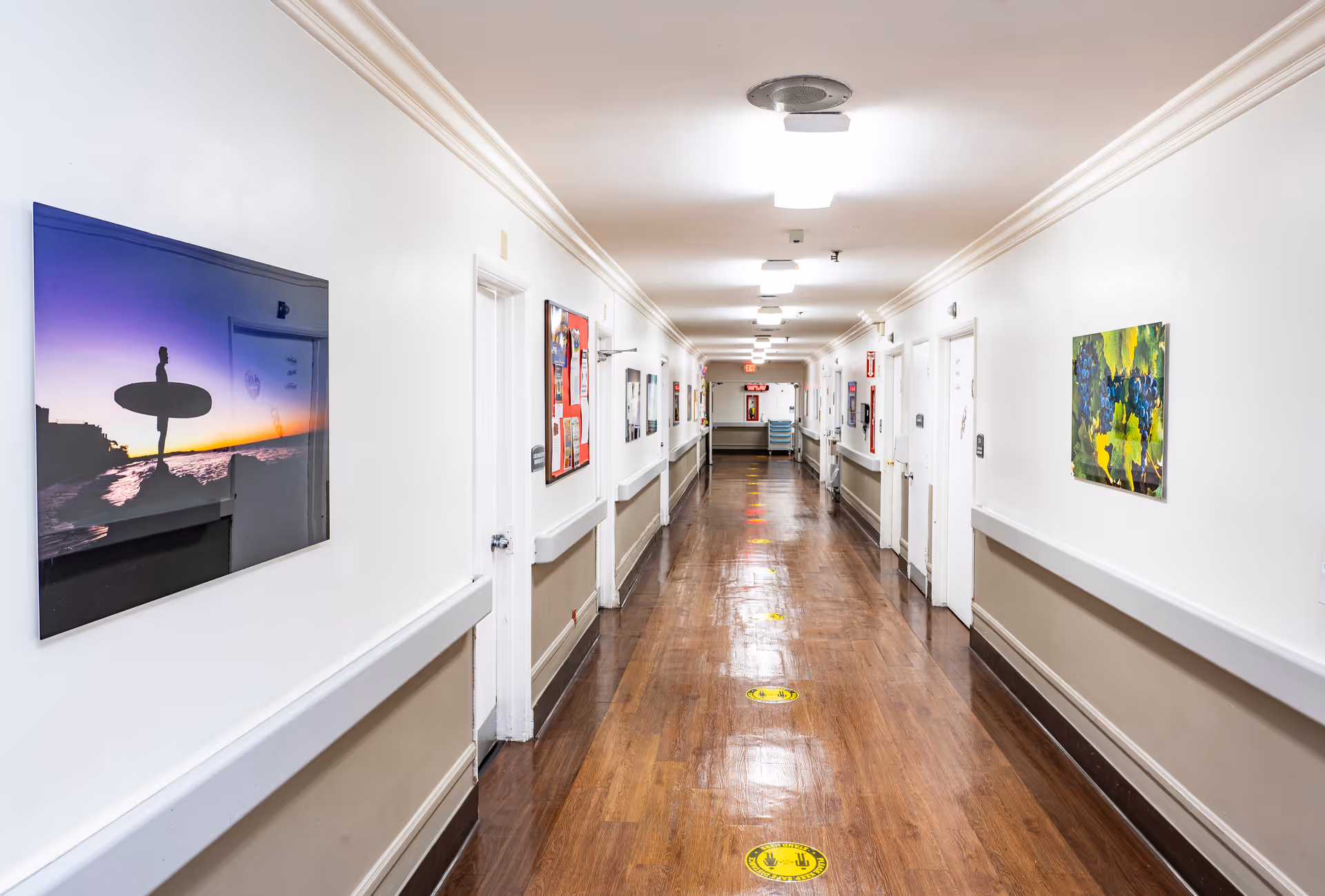 A long, well-lit hallway in a care facility with wooden floors and white walls. The walls are decorated with colorful artwork, including a picture of a surfer at sunset and a painting of grapes. Several white doors line both sides of the hallway, and there are handrails along the walls. Yellow social distancing floor markers are spaced along the floor.