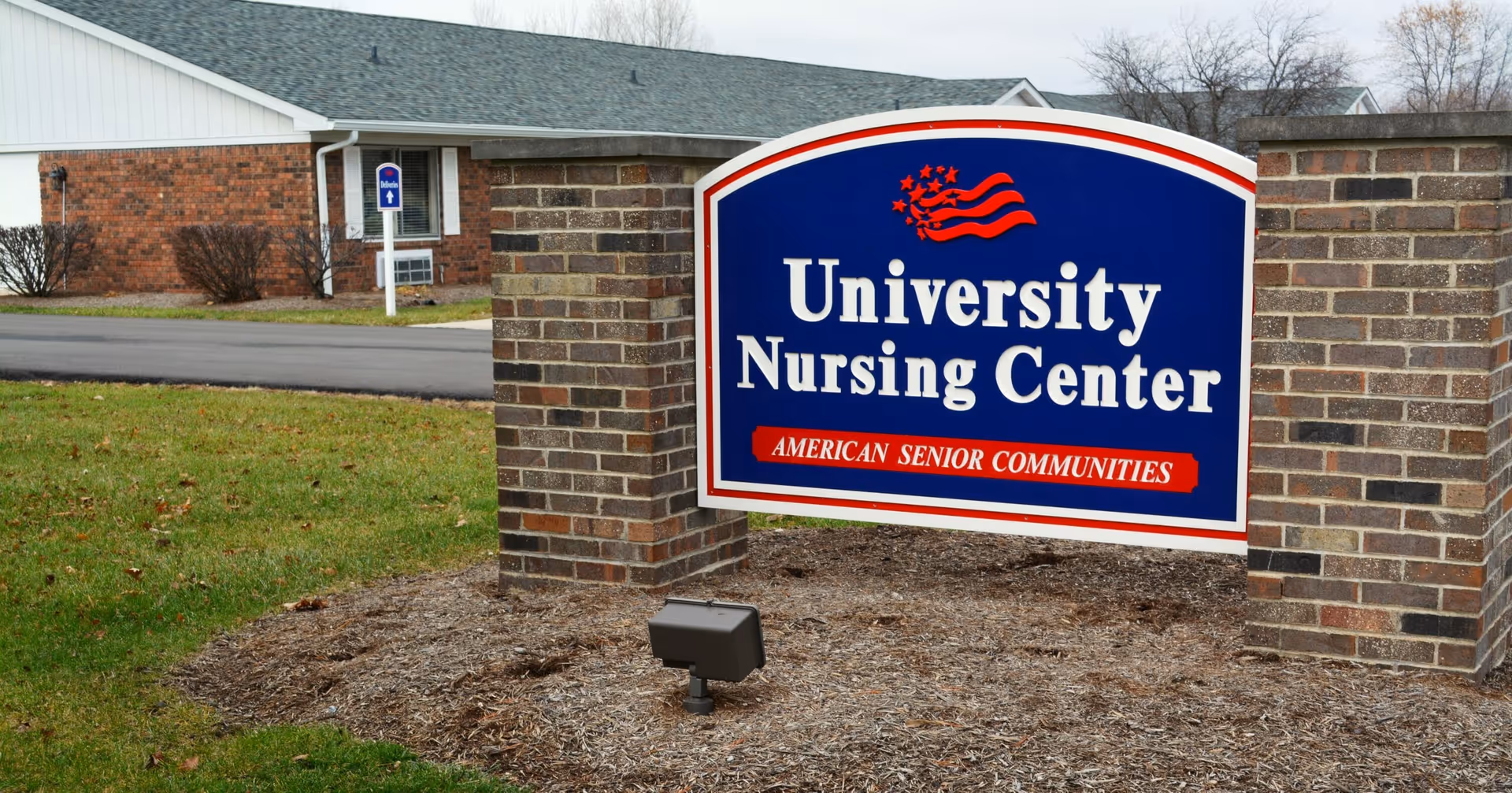 A large blue and white sign with red accents reading 'University Nursing Center' and 'American Senior Communities' in front of a brick building with a gray roof and a small driveway.