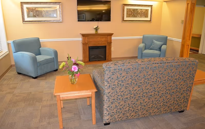 A cozy living room area with two blue armchairs, a patterned sofa, a wooden coffee table with a vase of flowers, a wooden electric fireplace, and a wall-mounted flat-screen TV. The walls are beige with white trim, and two framed artworks hang on the wall above the fireplace.