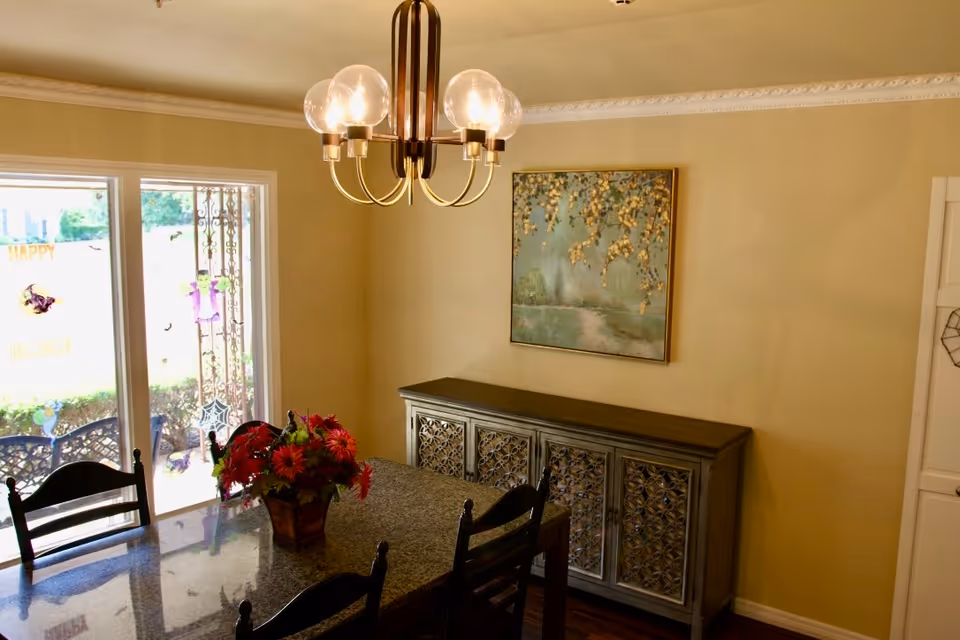 Dining room with a dark wooden table and chairs, a vase with red flowers on the table, a decorative sideboard with intricate patterns on the doors, a painting of a tree with golden leaves on the wall, a modern chandelier with five round bulbs, and sliding glass doors leading to an outdoor area decorated with Halloween-themed stickers.