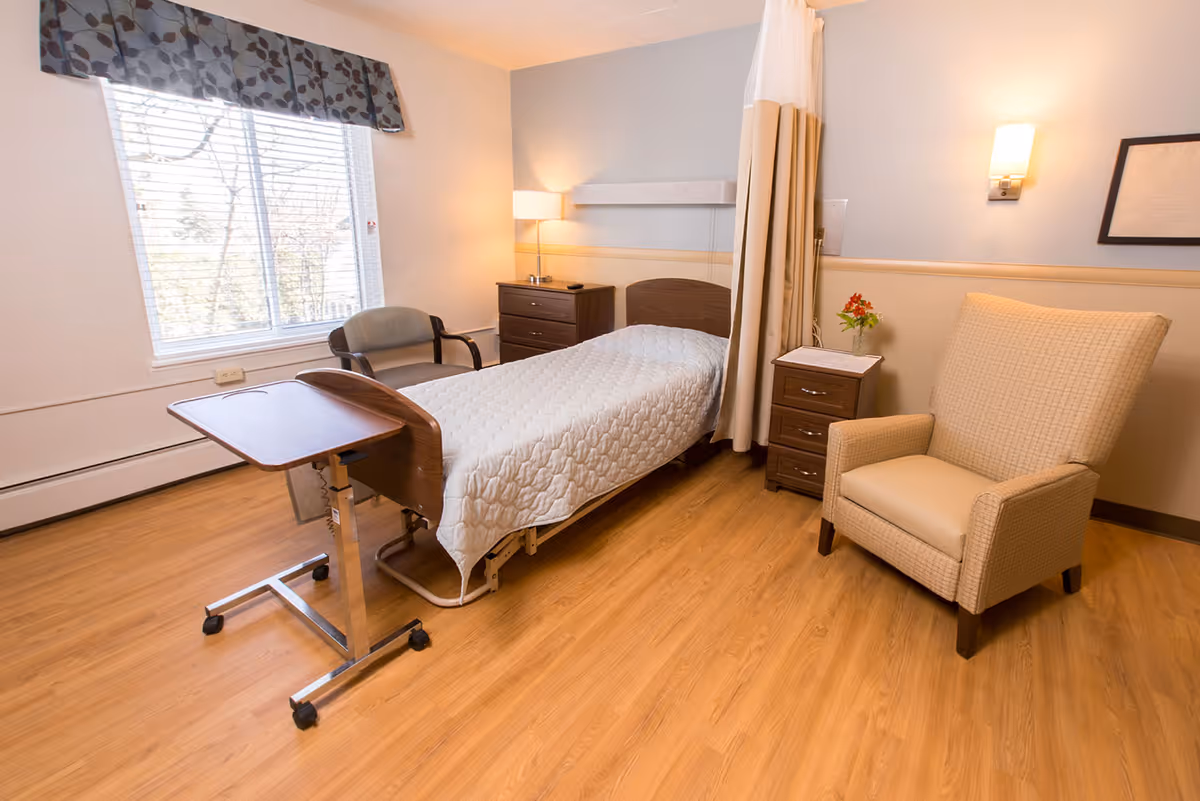 A well-lit senior care facility bedroom with a single hospital-style bed covered with a white quilted blanket. Next to the bed is a wooden bedside table with a small vase of flowers and a lamp on a dresser behind the bed. There is a beige upholstered armchair to the right and a chair near a window with blinds and a patterned valance. A mobile overbed table is positioned in front of the bed. The room has wood flooring and soft, neutral-colored walls.