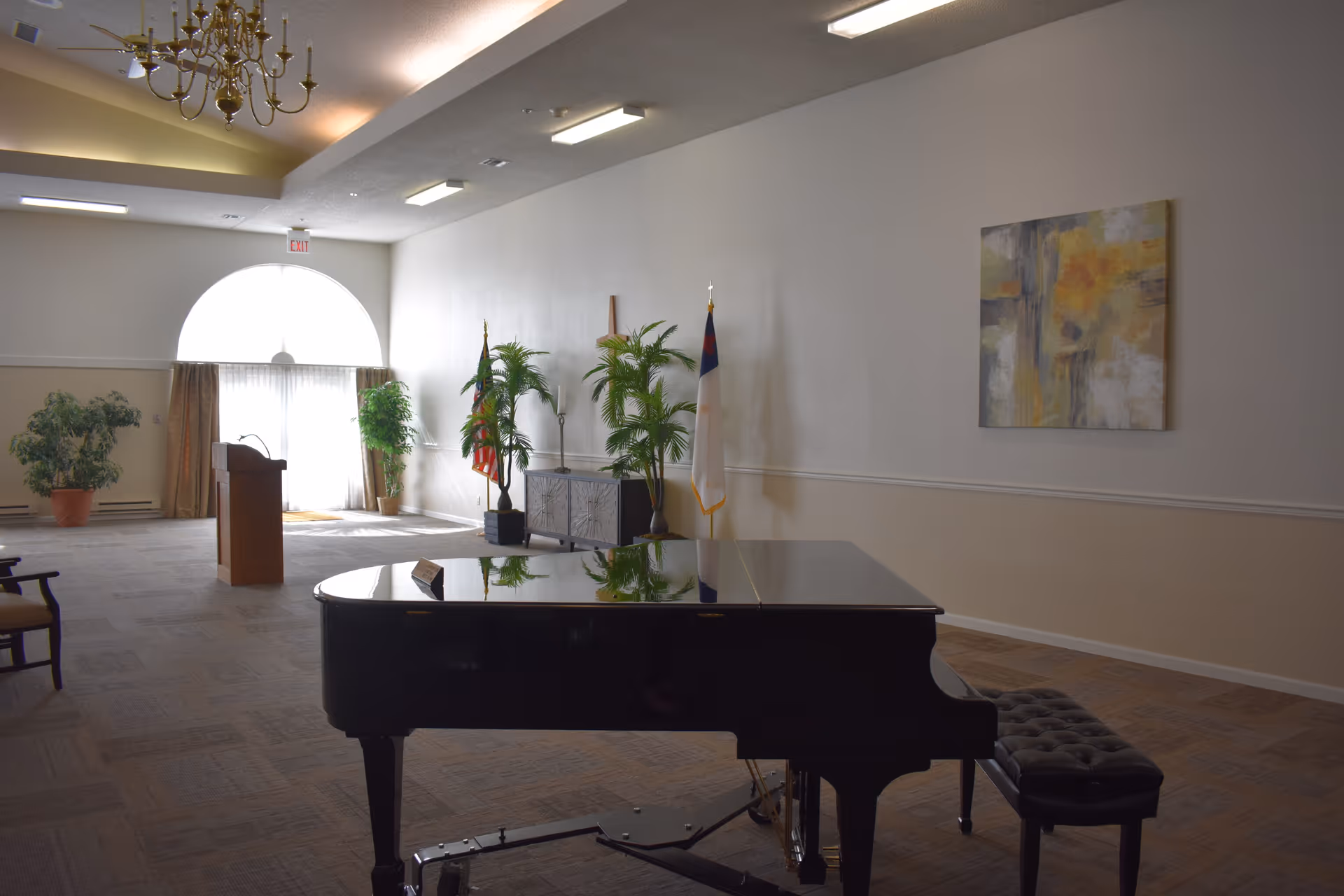A bright multipurpose interior room with a grand piano, podium, flags, potted plants and a chandelier.