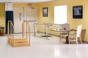 A bright therapy room with parallel walking bars, practice stairs, a table and chairs against yellow walls and an exit door.