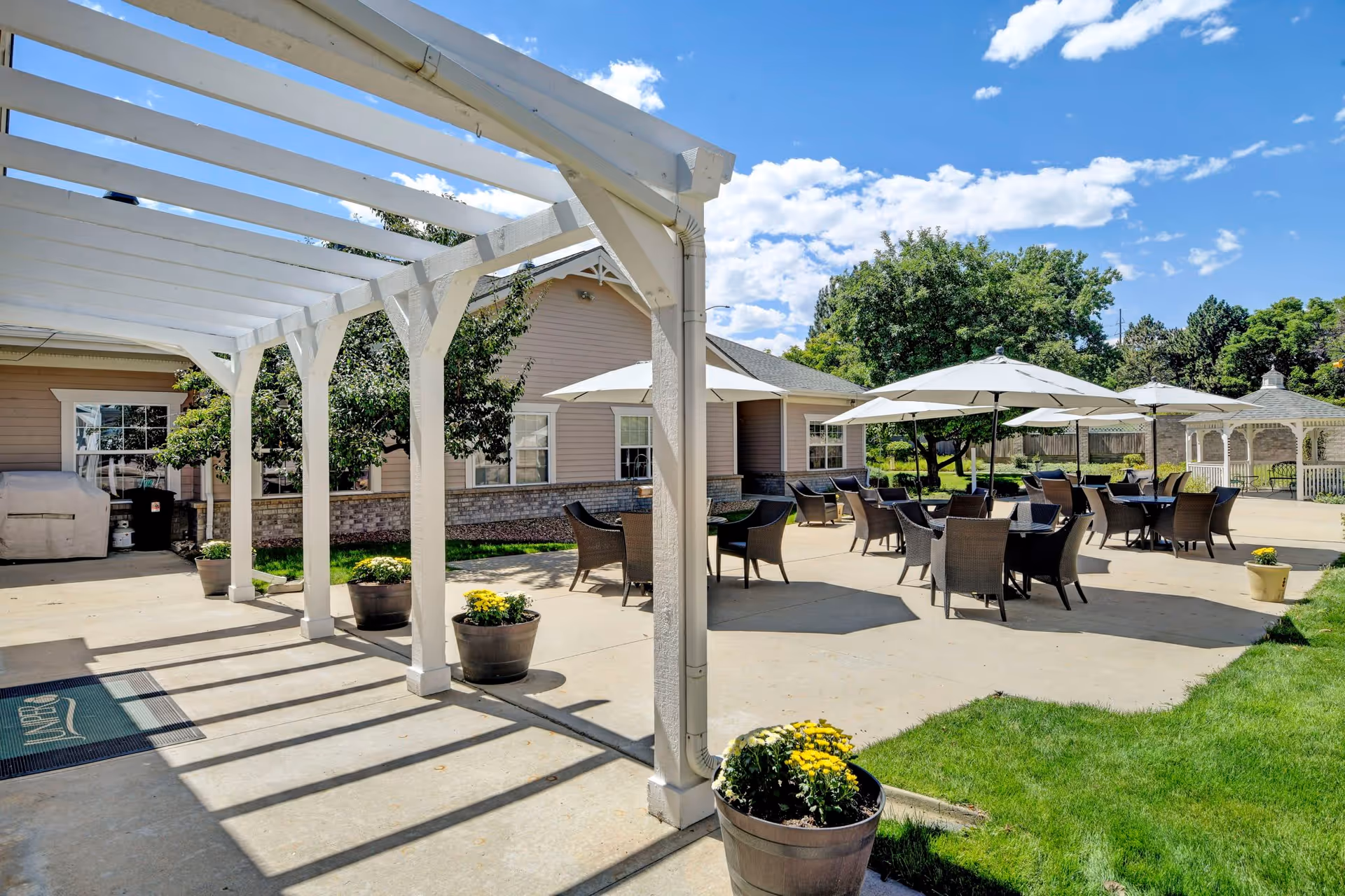 Outdoor patio area at a senior living facility with multiple tables and chairs under large white umbrellas, a white pergola casting shadows on the concrete ground, potted yellow flowers, green grass, trees, and a gazebo in the background under a blue sky with scattered clouds.
