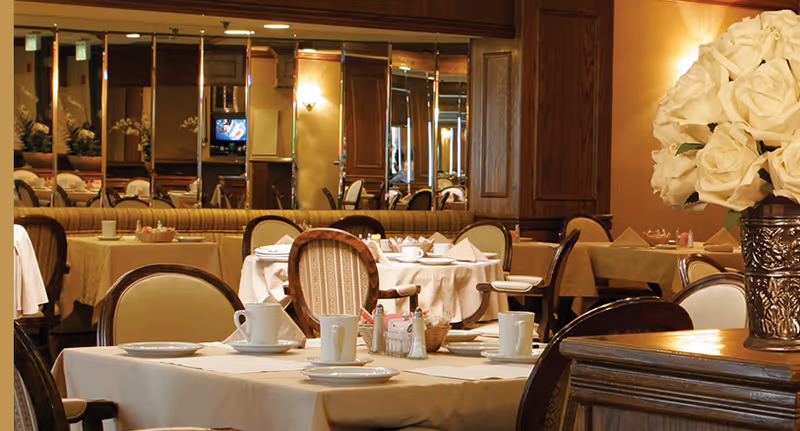 A warmly lit dining room with tables covered in white tablecloths, set with white cups, plates, and napkins. The room features wooden paneling, a large mirror on one wall reflecting the tables and chairs, and a vase with white roses on a wooden surface in the foreground.