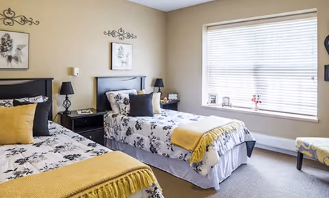 A bright bedroom with two twin beds featuring black and white floral bedding and yellow throws. Each bed has a black headboard and is accompanied by a black nightstand with a lamp. The room has beige walls with decorative wall art above each bed, a large window with blinds, and a patterned chair near the window.