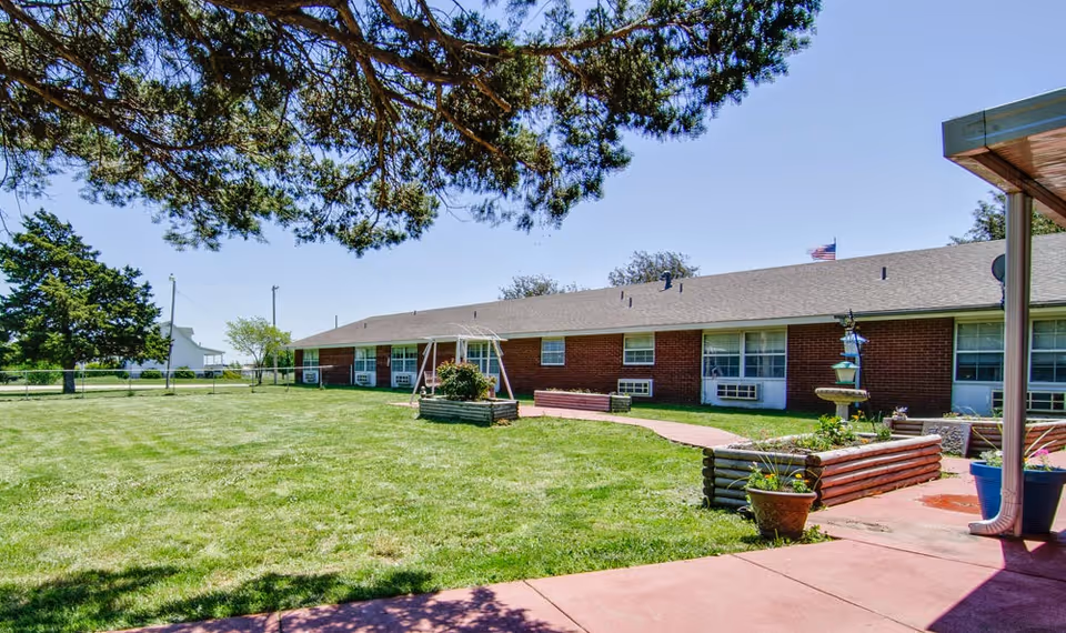 A single-story brick building with multiple windows and air conditioning units, surrounded by a well-maintained grassy lawn with a swing set, flower beds, and a paved walkway. Large trees provide shade in the foreground under a clear blue sky.