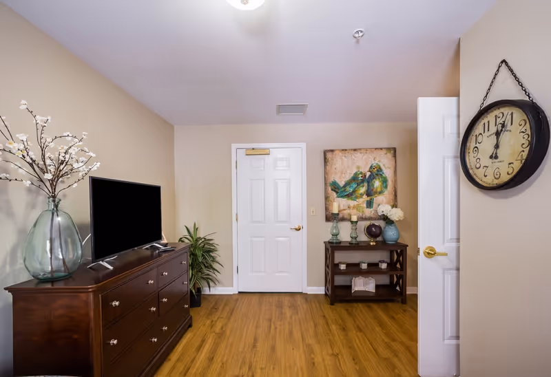 Interior living room with a wooden dresser and TV on the left, a console table with decor and artwork by a white door in the center, and a hanging clock on the right.