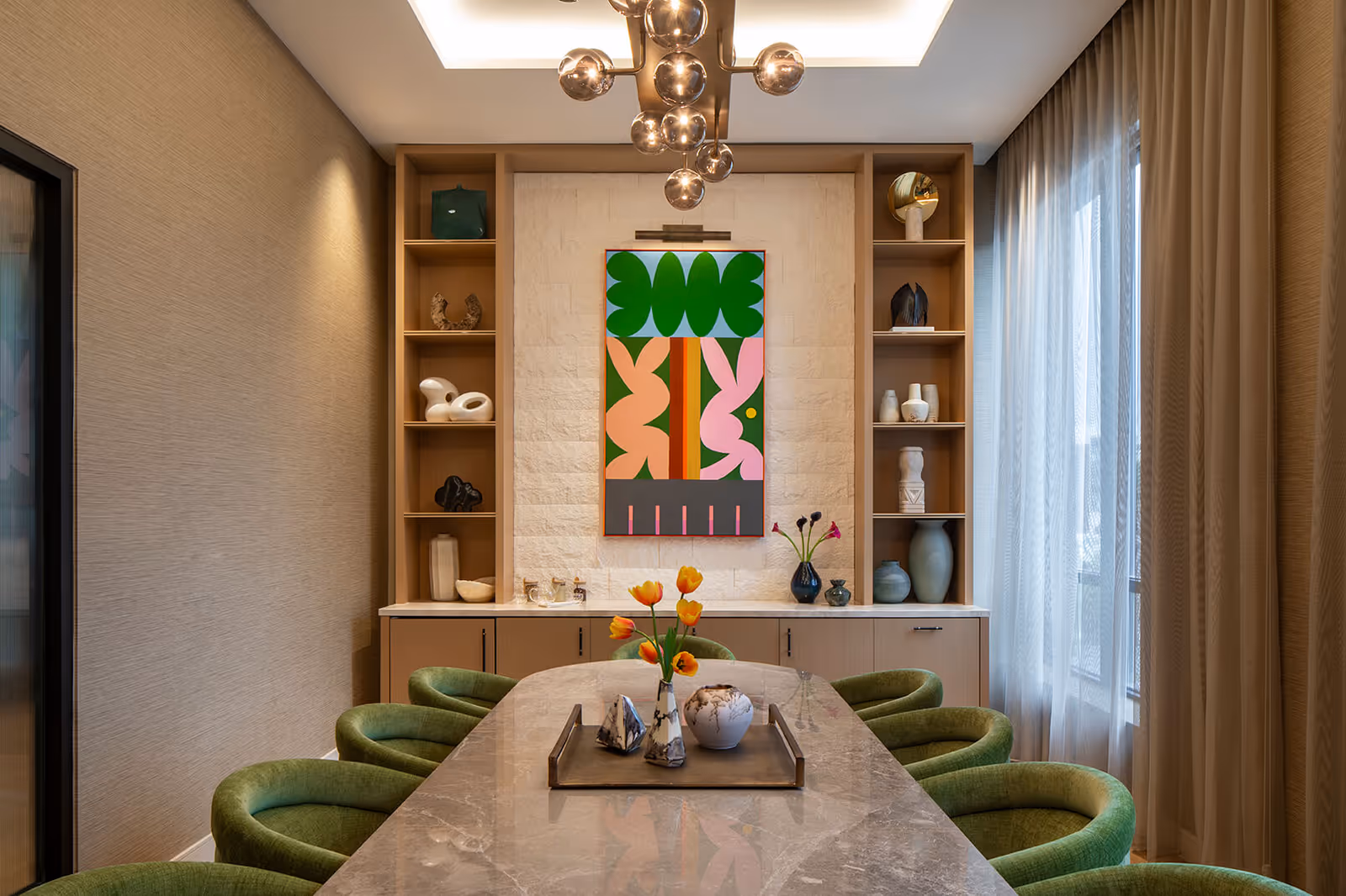 A modern dining room with a long marble table surrounded by green upholstered chairs. On the table is a tray holding decorative vases with orange tulips. The back wall features built-in wooden shelves with various decorative objects and a colorful abstract painting in the center. Large windows with sheer curtains allow natural light to fill the room.