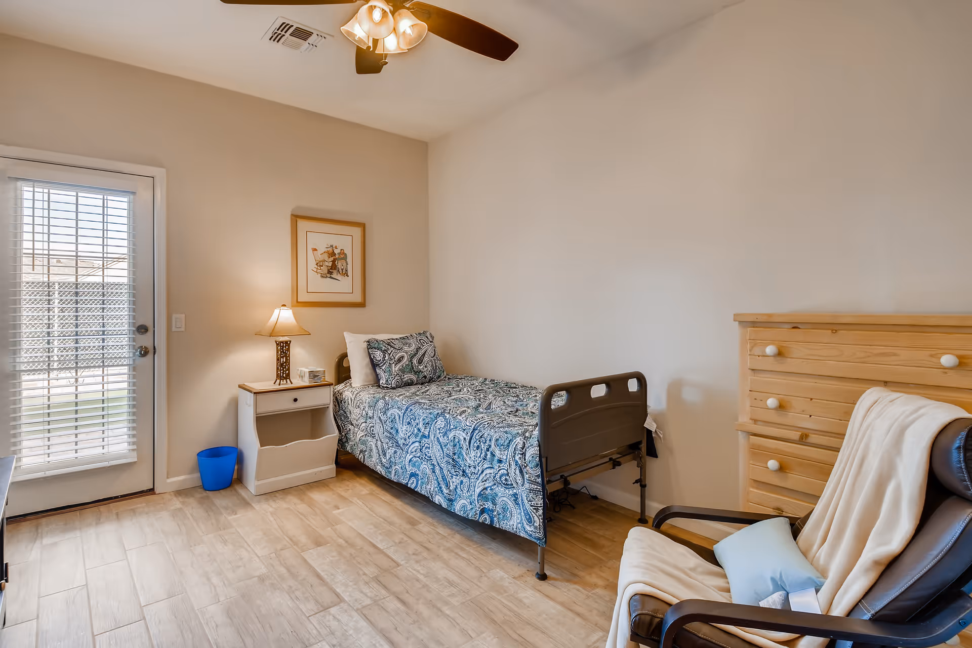 A small, neatly arranged bedroom in an assisted living facility featuring a single bed with a blue patterned bedspread, a wooden nightstand with a lamp and clock, a wooden dresser, and a comfortable chair with a blanket and pillow. A door with blinds leads outside, and a ceiling fan with lights is mounted above.