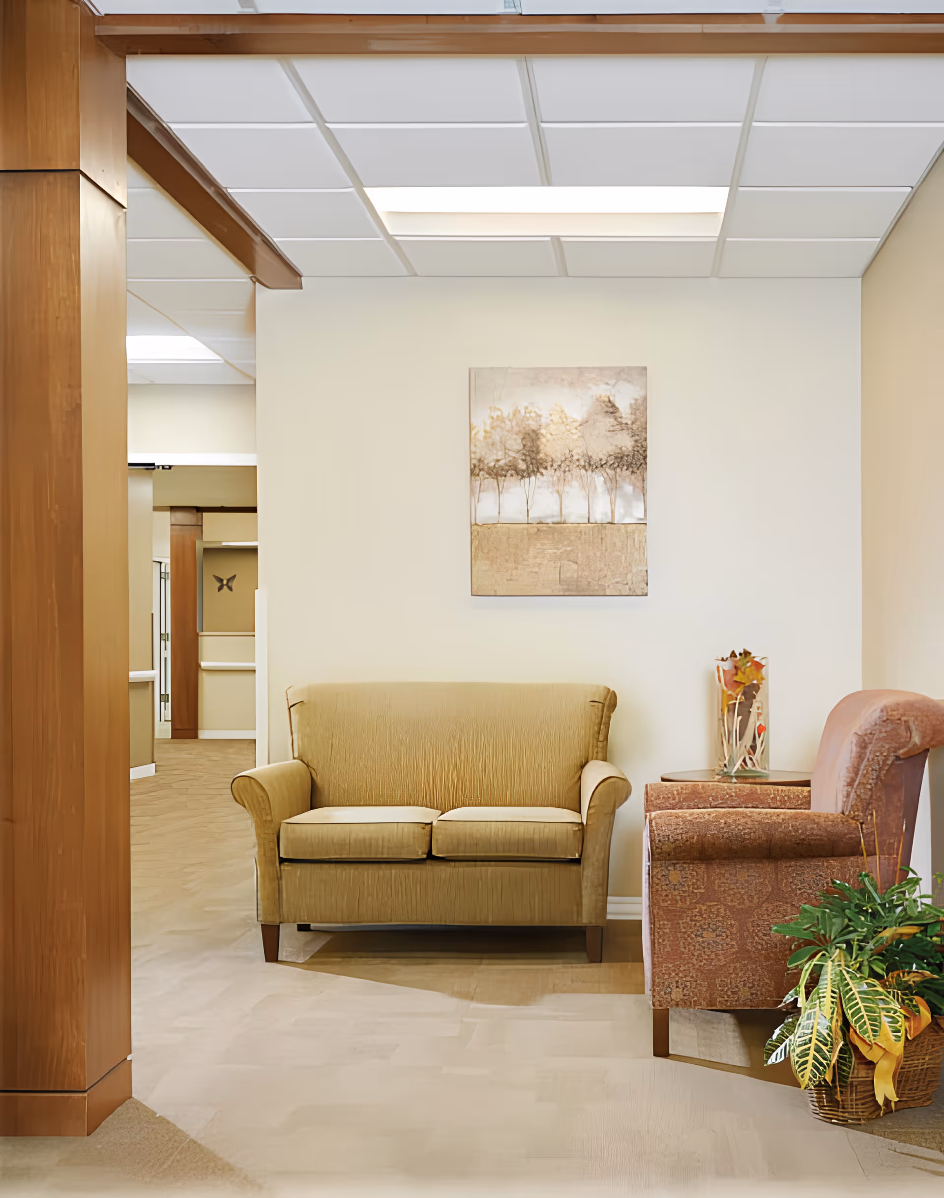 A cozy seating area in a senior living facility with a beige loveseat and a patterned armchair. A small side table with a decorative vase and a potted plant are next to the armchair. A painting of trees hangs on the wall above the loveseat. The space has a neutral color palette and a soft carpeted floor.
