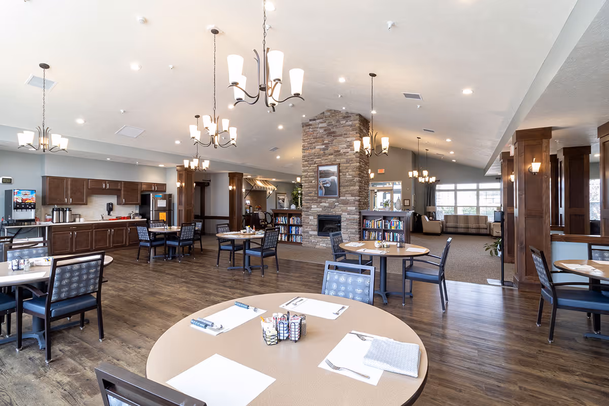 A spacious dining area in an assisted living facility with round tables set with placemats, utensils, and napkins. The room features wood flooring, multiple chandeliers, a stone fireplace with bookshelves on either side, and a kitchen area with cabinets and a beverage dispenser in the background. Large windows allow natural light to fill the space.