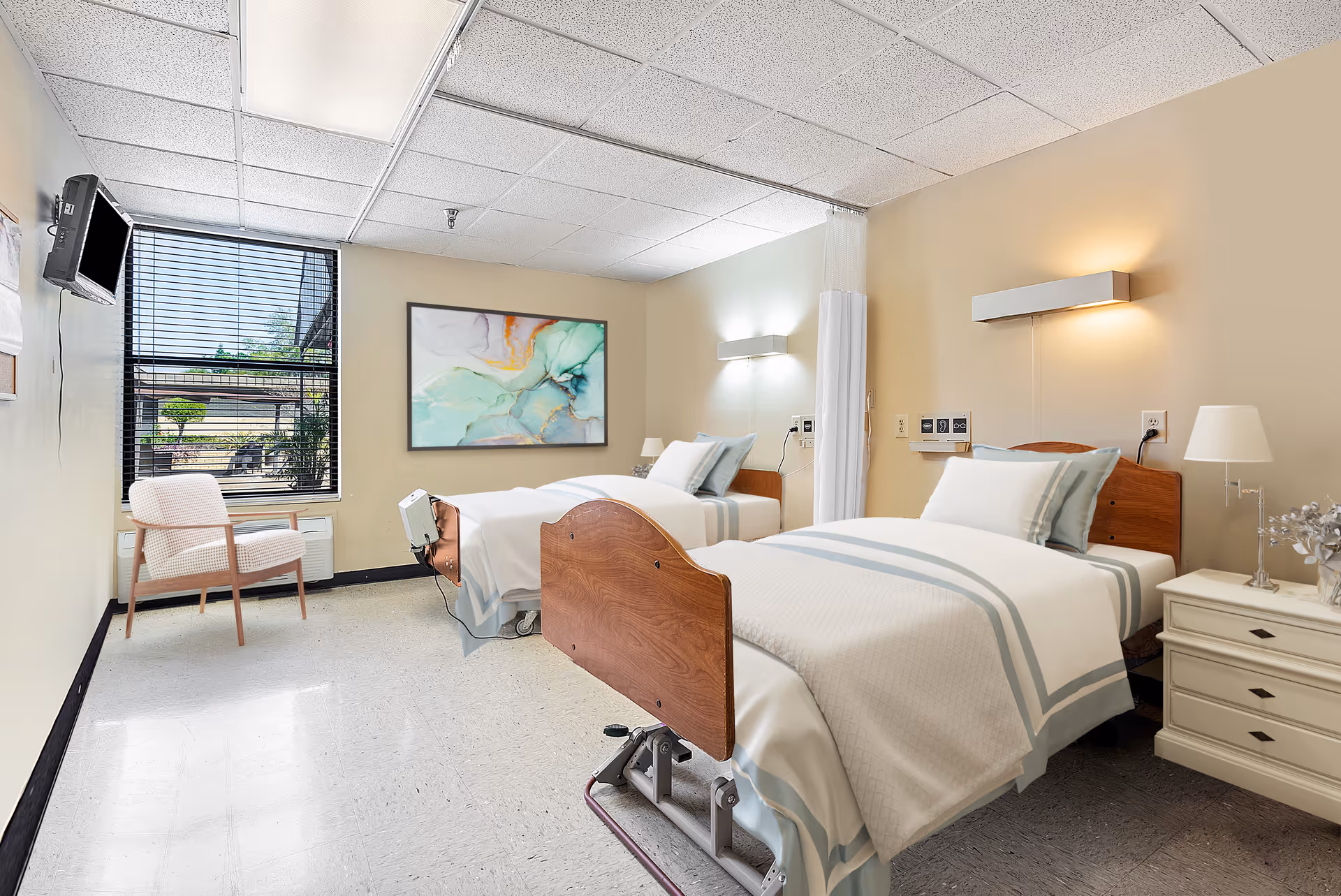 A bright and clean nursing facility room with two hospital beds made up with white and light blue bedding. Each bed has a wooden headboard and is positioned against a beige wall with mounted lights above. A white nightstand with a lamp and decorative item is placed between the beds. A window with black blinds lets in natural light, and a cushioned chair is positioned near the window. A colorful abstract painting hangs on the wall between the beds and the window.