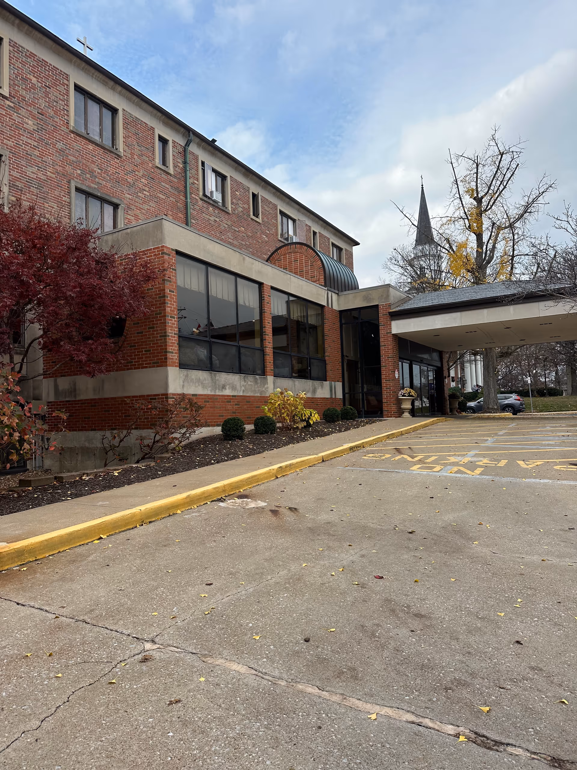 Exterior view of a brick building with large windows and a covered entrance. There is a cross on the top left corner of the building, indicating it may be affiliated with a church. The parking area in front has yellow painted curbs and markings. Trees with autumn foliage are visible around the building under a partly cloudy sky.