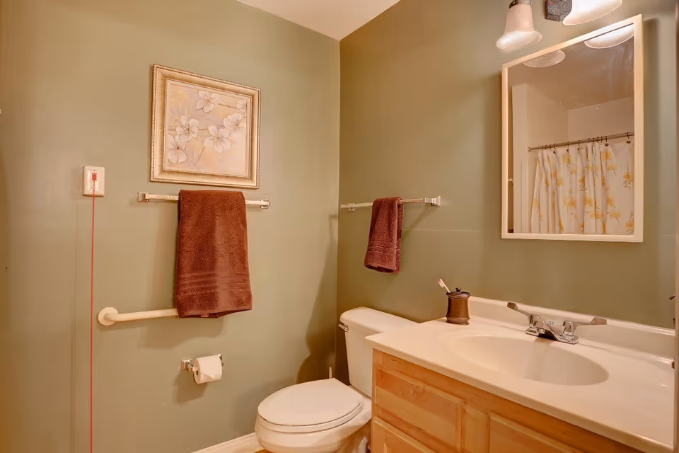 A bathroom with olive green walls featuring a toilet, a wooden vanity with a white sink, a mirror above the sink, two brown towels hanging on towel racks, a floral shower curtain, a framed floral artwork on the wall, and a grab bar next to the toilet.