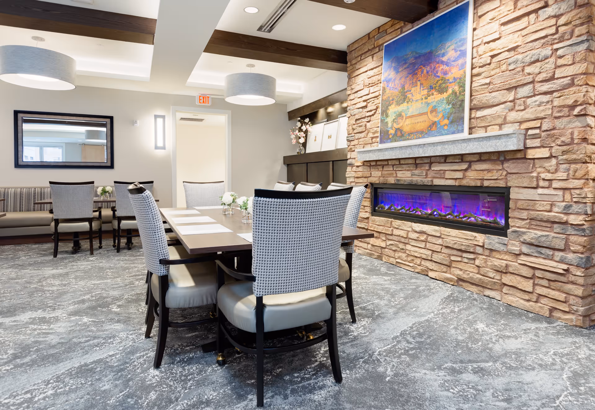 A bright dining area with a table and upholstered chairs in front of a stone wall with a built-in electric fireplace and framed artwork.