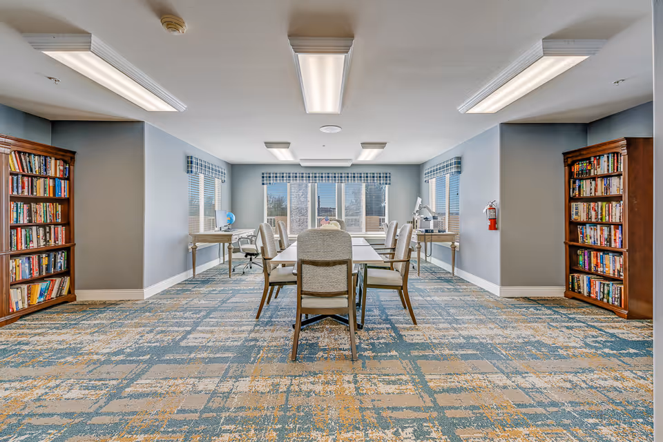 Bright communal activity room with a central table and chairs, bookshelves on both sides, and large windows at the far end.