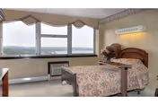 A furnished senior bedroom with a single bed, nightstand with flowers, armchair, and large windows overlooking the landscape.