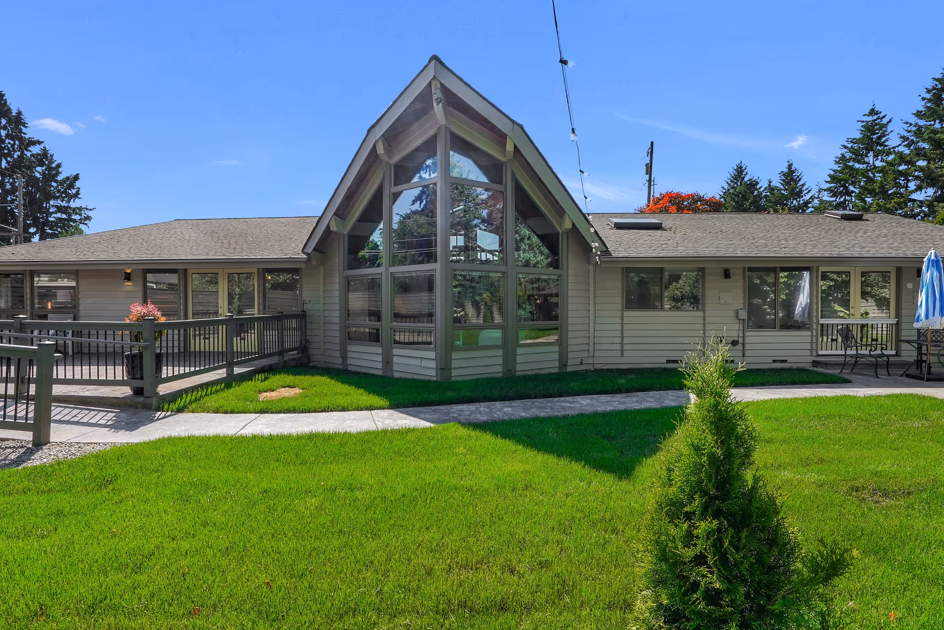 Exterior view of a single-story building with large triangular windows in the center, surrounded by a well-maintained green lawn and a paved walkway. The building has light-colored siding and a sloped roof with string lights hanging above the walkway. There is outdoor seating with umbrellas on the right side.