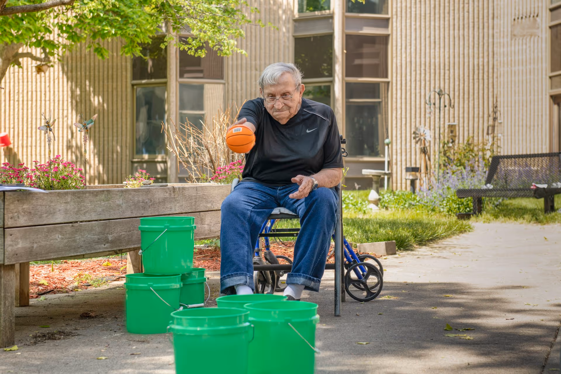 An elderly man sitting outdoors on a chair next to a walker, playing a game where he throws an orange ball towards a group of green buckets arranged on the ground. The setting is a garden area with flowers, a wooden bench, and a building with large windows in the background.