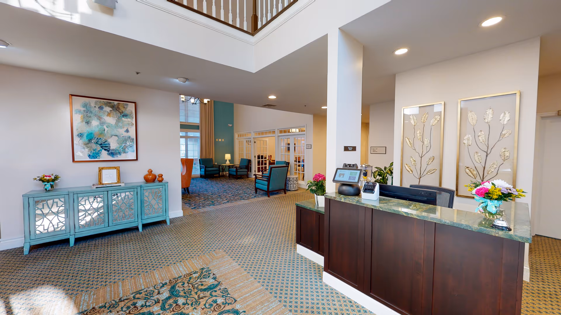 Reception area of a senior living facility with a dark wood front desk topped with green marble, decorated with flowers and framed leaf artwork on the wall behind. To the left, a turquoise cabinet with decorative mirrored doors holds vases and a framed item, with an abstract painting hanging above. In the background, a seating area with blue armchairs and a lamp is visible through glass-paneled doors.