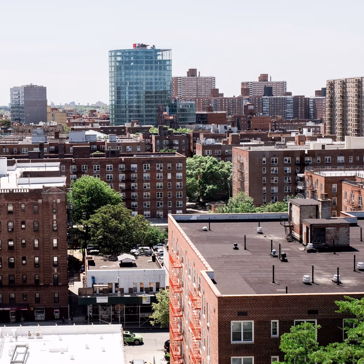 High-angle view of a city neighborhood with multiple brick apartment buildings, a rooftop in the foreground, and a glass cylindrical tower in the distance.