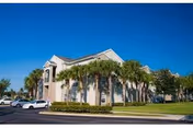 Front view of a two-story beige apartment building with palm trees, parked cars, and a clear blue sky.