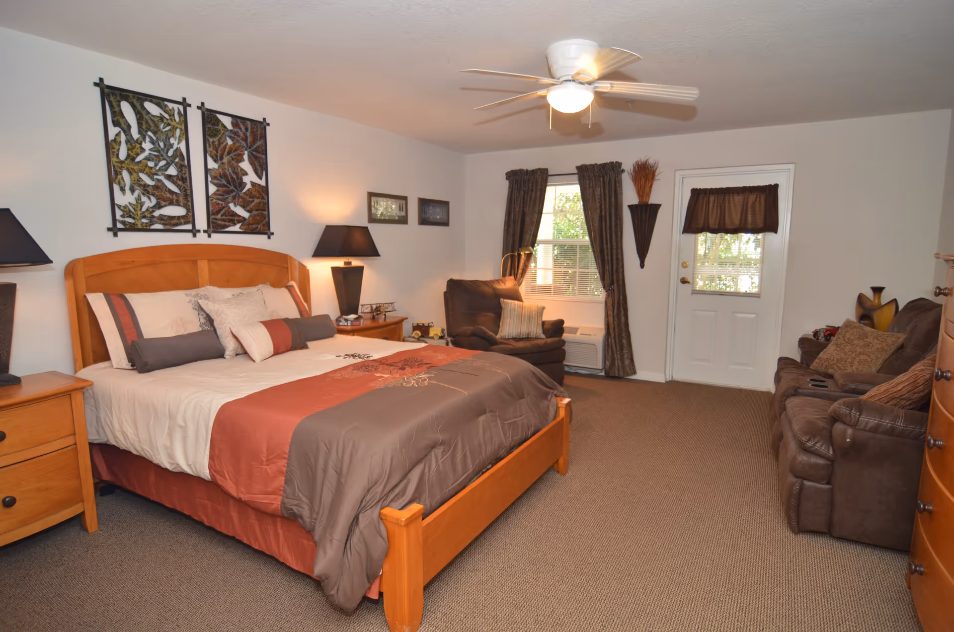 A cozy bedroom with a wooden bed frame and a neatly made bed with brown and rust-colored bedding. There are two wooden nightstands with black lamps on either side of the bed. The room also features a brown recliner chair near a window with dark curtains, a white door with a small window covered by a brown valance, and a brown sofa with cushions. The walls are decorated with framed artwork and a decorative wall piece. The ceiling has a white fan with a light.