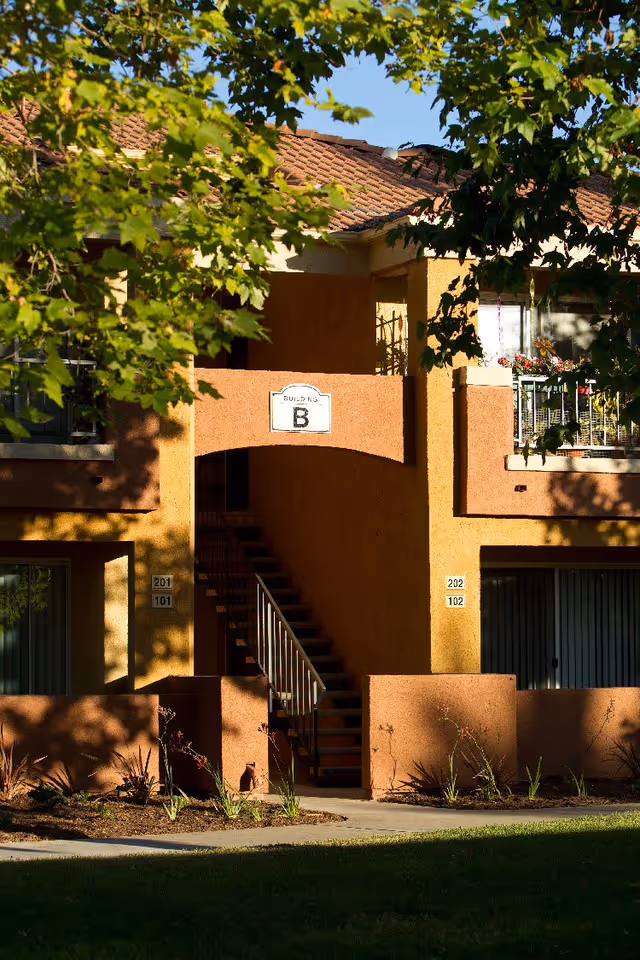 Exterior view of a two-story residential building labeled Building B with stair access to the upper floor, surrounded by trees and landscaping in the foreground.