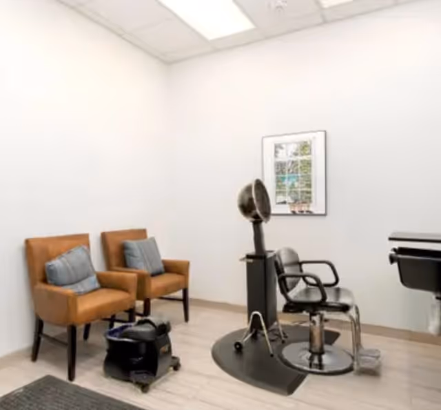 A small salon room with two brown armchairs with blue cushions, a black salon chair with a hair dryer hood, a black footrest, and a framed picture on the white wall.