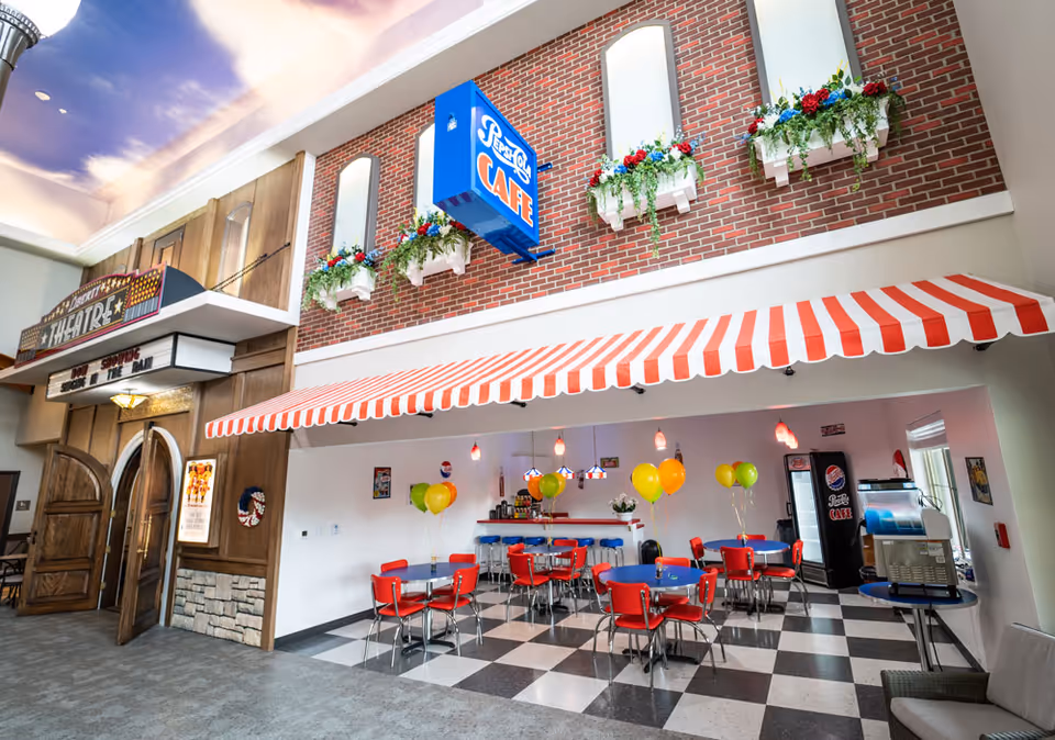 Interior view of a retro-style cafe area with red and white striped awning, red chairs, blue tables, and colorful balloons. The cafe is decorated with flower boxes on a brick wall above and a blue sign that reads 'Pepsi Cola Cafe'. To the left, there is a wooden entrance to a theater with a marquee sign.
