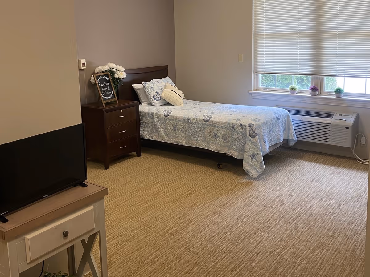 A simple, clean bedroom with a single bed covered in a light blue quilt with seashell patterns. Next to the bed is a dark wooden nightstand with a vase of white flowers and a small framed sign. A window with blinds and three small potted plants on the windowsill lets in natural light. A small TV sits on a light-colored wooden table in the foreground.